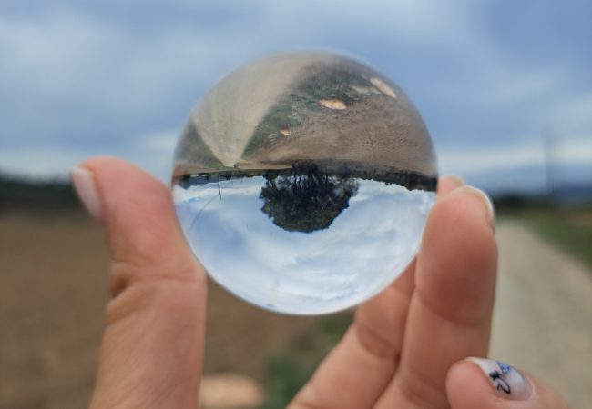 Mano de mujer sujetando una pequeña esfera de cristal, dentro de la esfera el pasisaje se ve invertido.